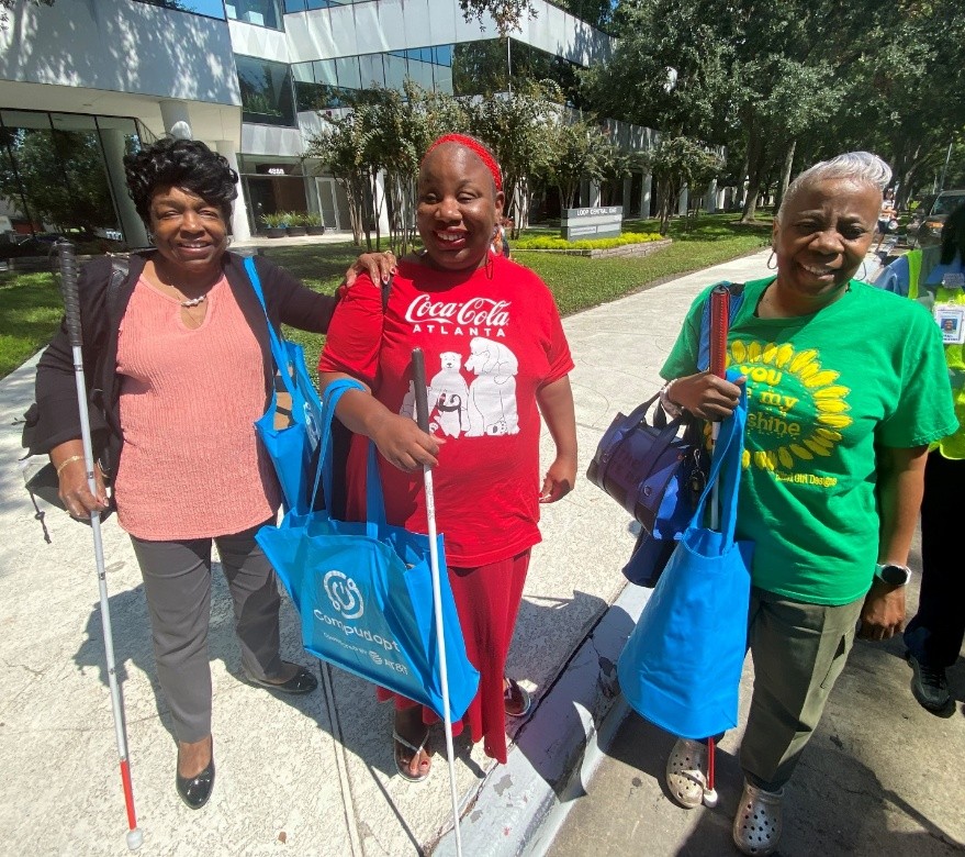Three Black women with white canes, lined up on a sunlit curb and smiling at the camera.