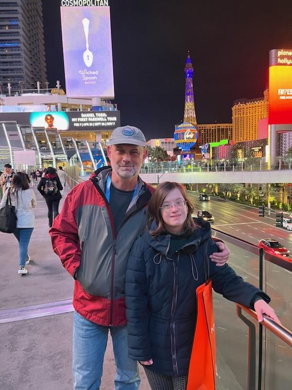 Michael and Sarah Brhlik posing against Las Vegas skyline at night. Michael (left) is wearing a gray baseball cap, a red-and-gray windbreaker, and jeans; he has his left arm around Sarah's left shoulder. Sarah has long brown hair and is wearing glasses and a dark jacket; her left hand is on a silver-colored railing.