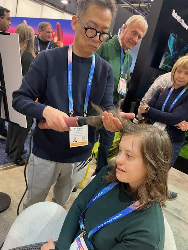 Sarah sitting in a white chair, having her hair curled with a pink-handled iron held by a man with short, curly hair and black-framed glasses.