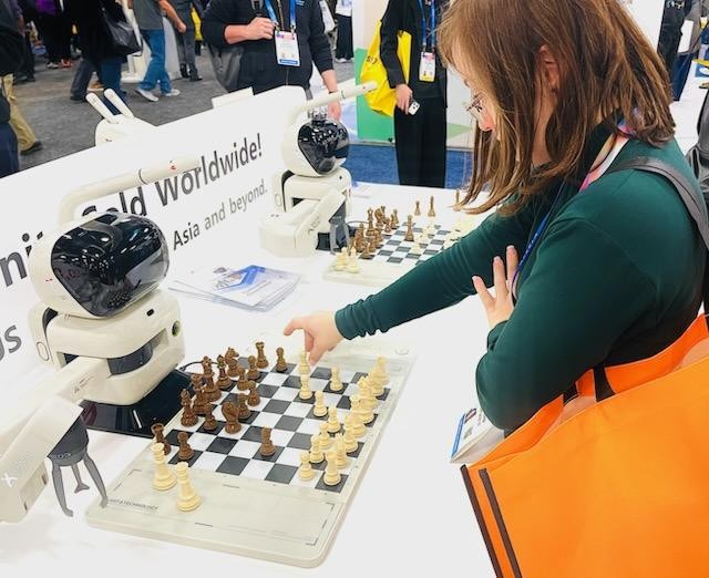 Sarah moving a white piece on a standard chessboard. Facing her on the other side of the board is a small, white robot with a black-visor face.