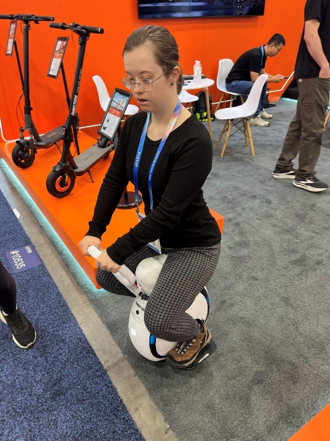 Sarah sitting on a white device that resembles a small wheel with a wagon handle. Two black stand-up scooters are behind her to the left; a white table and chairs are to the rear right. A man in a dark shirt is sitting in the rearmost chair; someone else, visible from the shoulders down, is standing to the right.