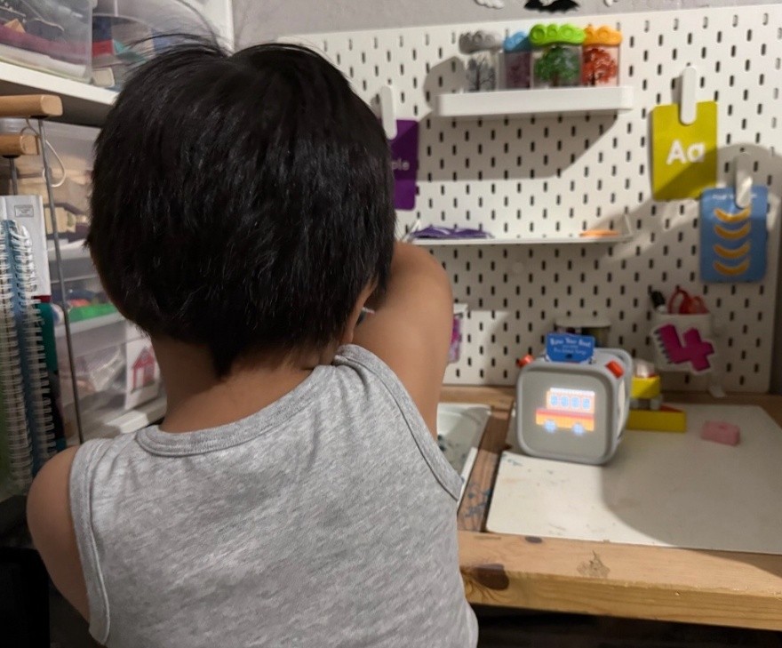 Child with short dark hair, back to camera, facing a work desk. Yoto player (gray box showing digital image of van) on desk to child's right.